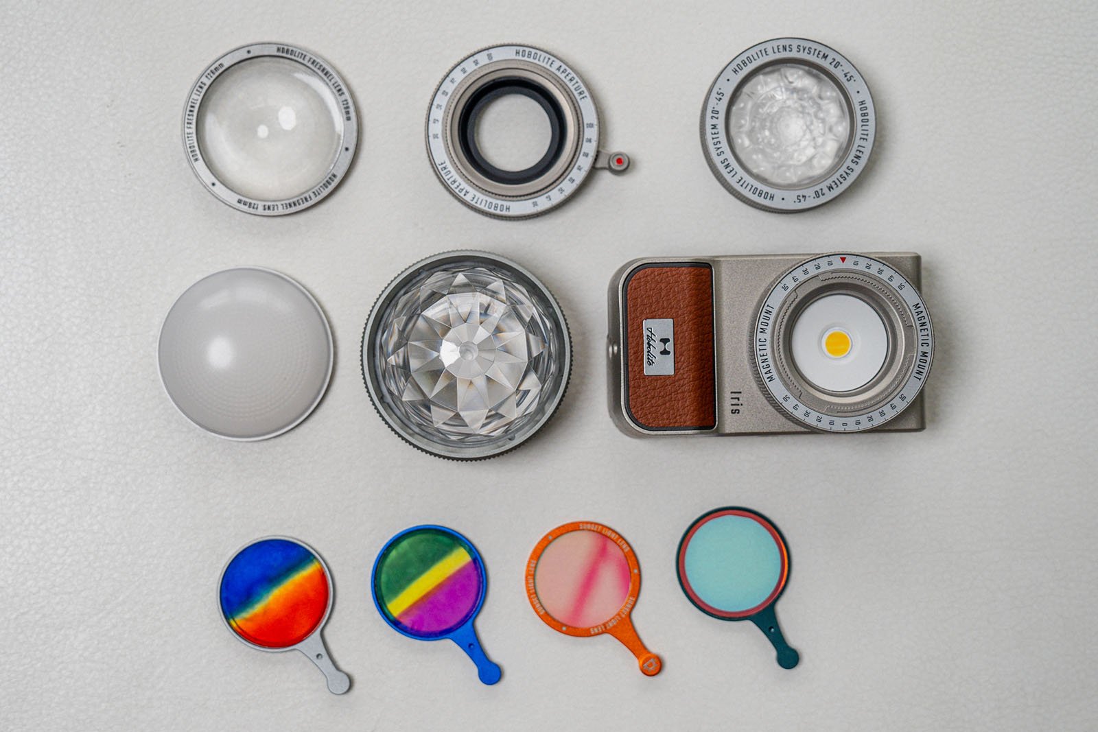 A collection of photography accessories arranged on a white surface, including camera filters, a lens, a camera body, and various colored lens covers. The colored covers include a blue, red, orange, and rainbow filter.