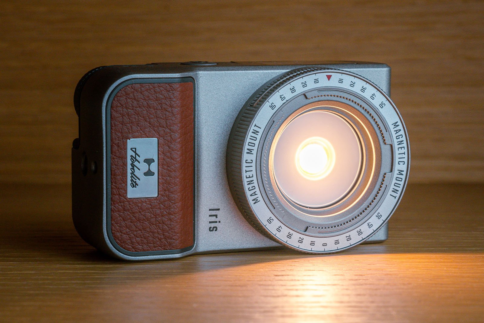 A retro-style camera with a prominent, illuminated lens, labeled "MAGNETIC MOUNT" around the lens ring. The camera has a silver body with a brown leather-like textured panel on the side, and is resting on a wooden surface.