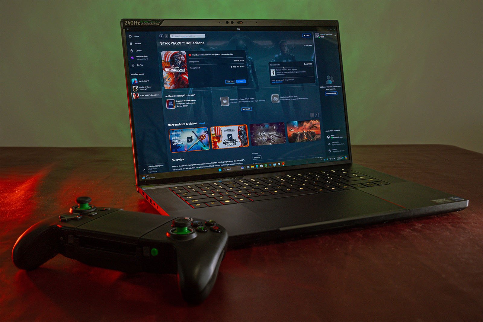 A gaming laptop with its screen displaying a game library is placed on a red surface. Next to the laptop is a wireless gaming controller. The background features a subtle gradient of green light.