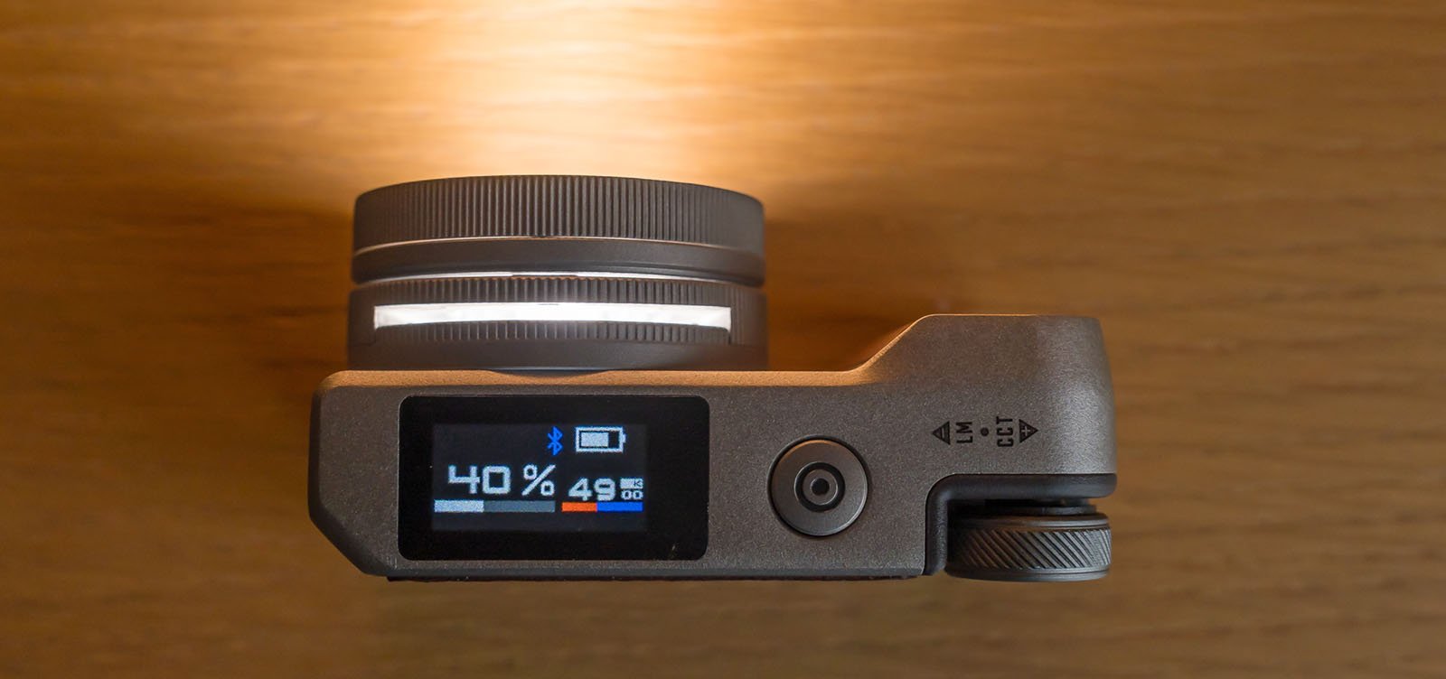 A top view of a digital camera placed on a wooden surface, showing battery information on its small screen. The screen displays a battery level of 40% and indicates the camera is on. The lens and control dial are visible at the left side of the camera.