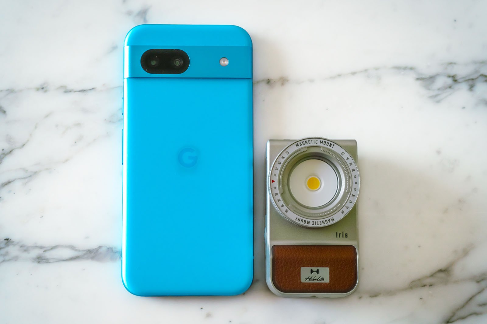 A cobalt blue smartphone with a "G" logo on the back lies on a white marble surface. Next to it is a compact, retro-styled camera with a magnetic mount and "Holstic" branding on a leather-textured grip.
