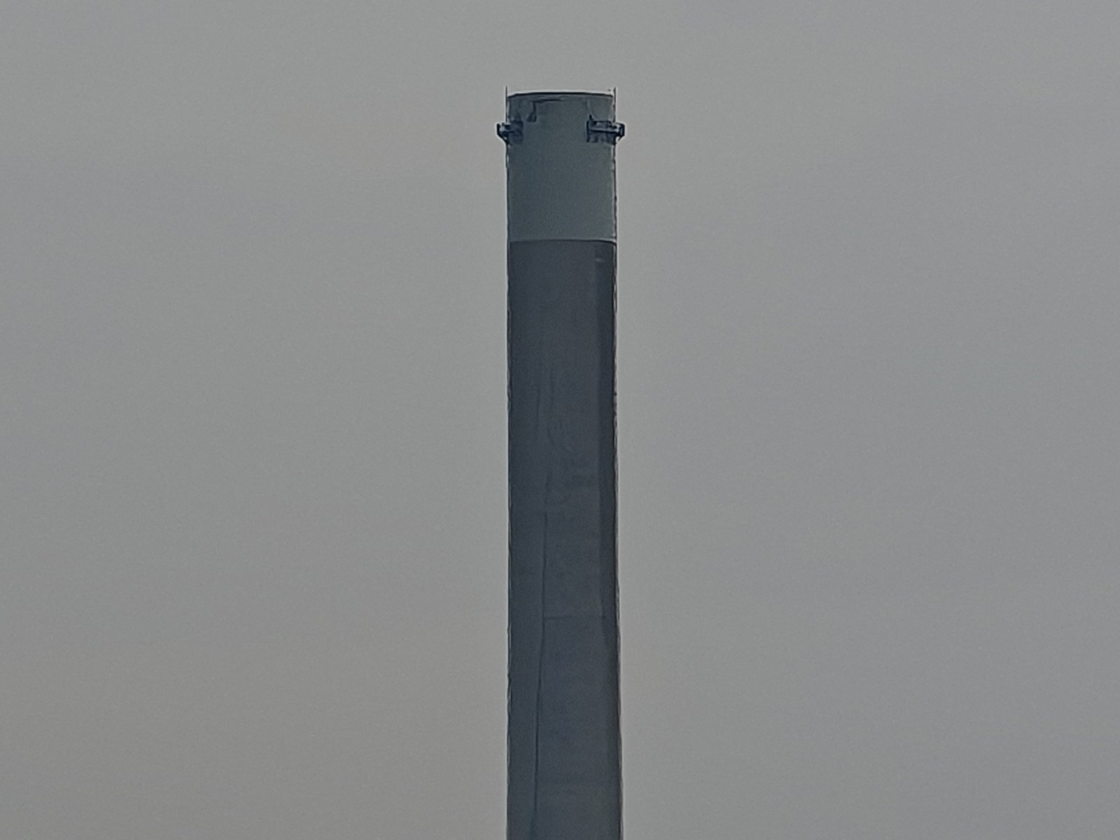 A close-up view of a tall, cylindrical industrial smokestack against a hazy gray sky. the stack has a slightly weathered surface and features two pipes protruding from its top.