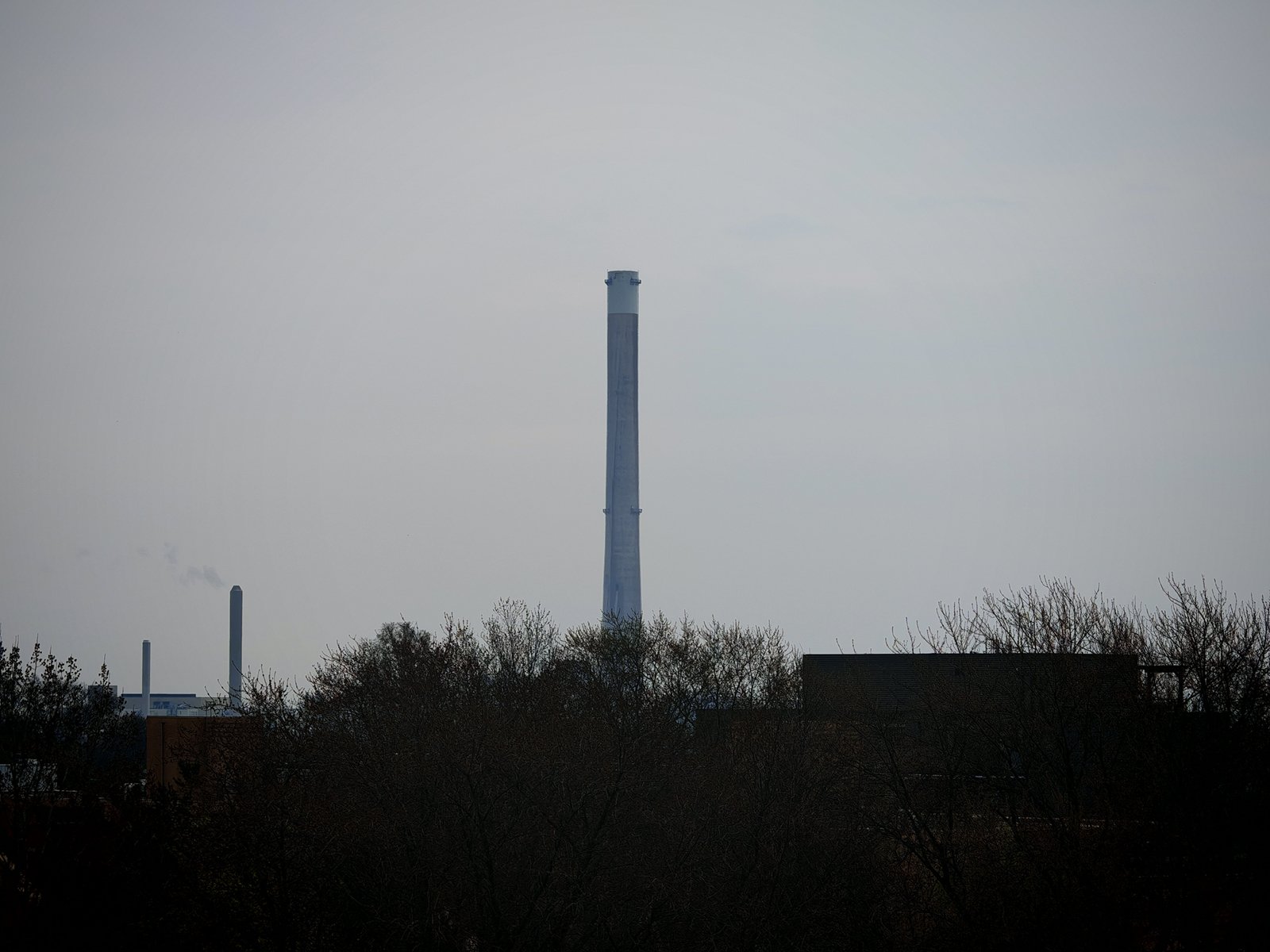 A silhouette of an industrial area with a tall chimney and several smaller structures, against a hazy sky.