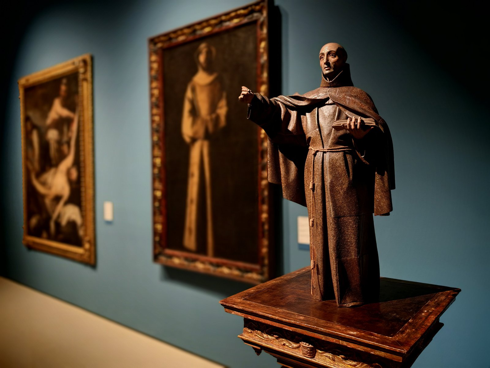 A wooden sculpture of a robed historical figure gesturing with one hand, displayed in a museum with paintings in the background. the lighting highlights the sculpture's detailed craftsmanship.