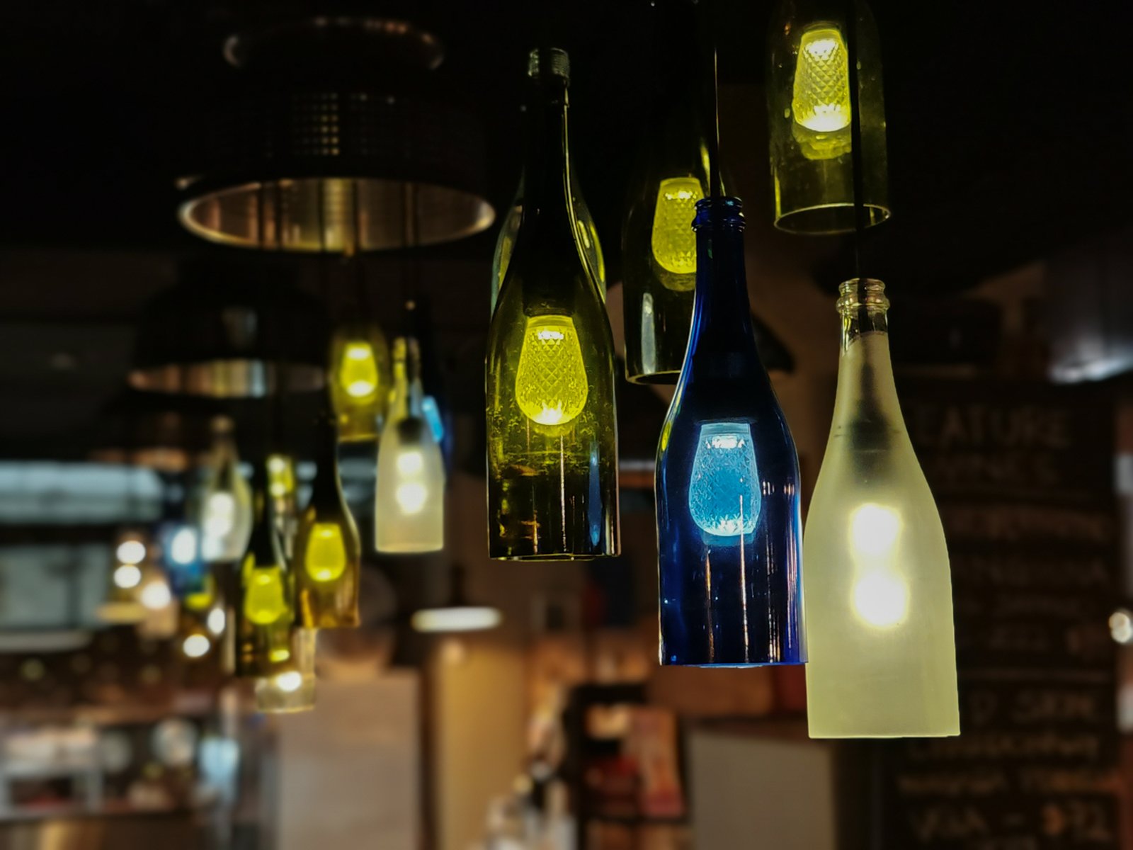 Decorative light fixtures made from recycled glass bottles in various colors, hanging in a dimly lit bar ambiance. the bottles emit a soft, warm glow, enhancing the cozy atmosphere.