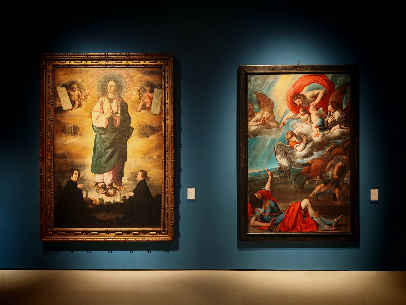 Two large framed paintings displayed on a dark blue wall in a gallery, one depicting a saint adorned in green with onlookers, and the other showing a dramatic religious scene with vibrant colors.