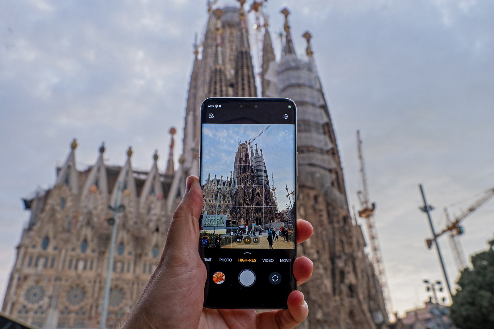 A hand holding a smartphone capturing a photo of the sagrada familia in barcelona, displayed on the phone screen, with the actual cathedral blurred in the background.
