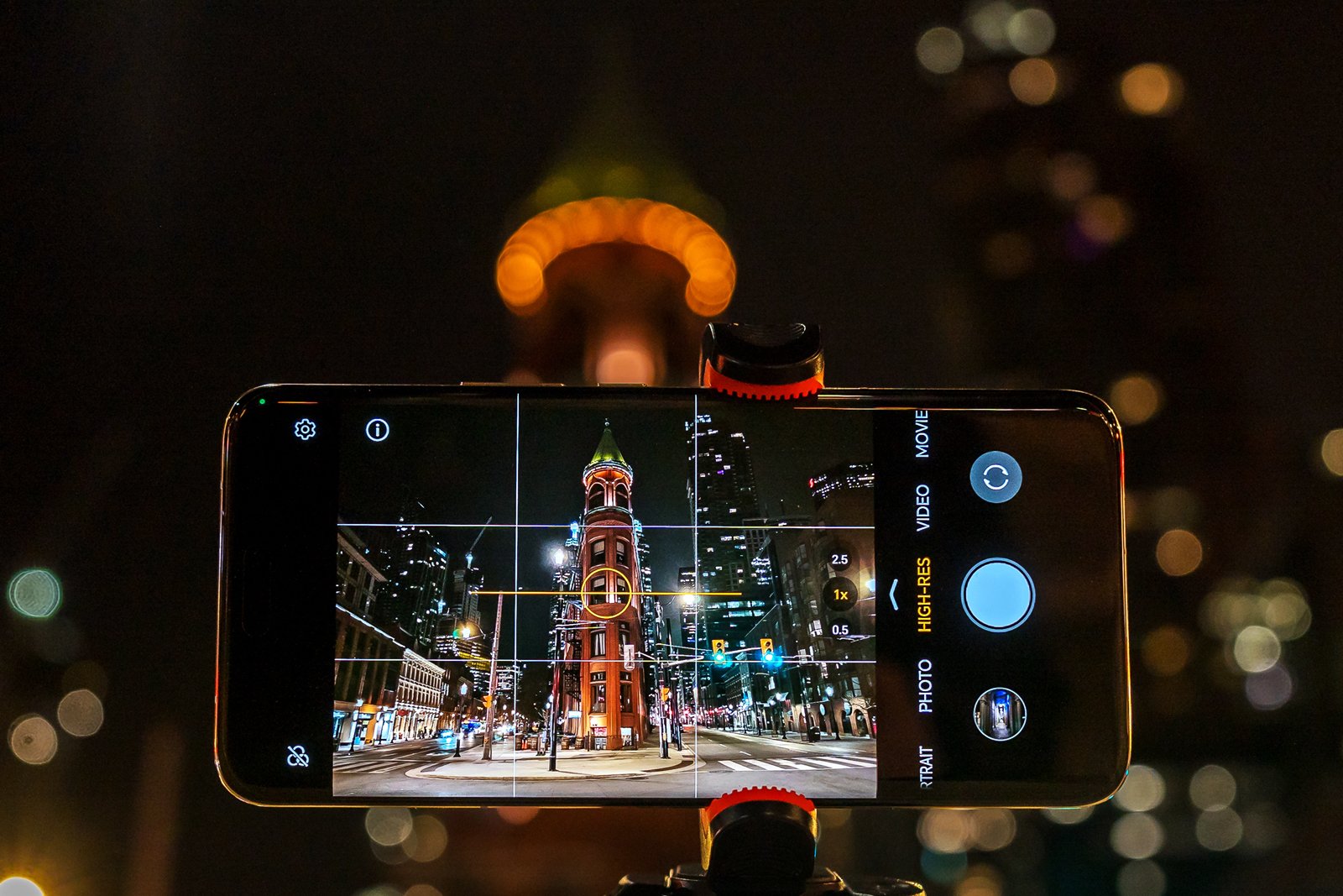 A smartphone screen displaying a city street at night framed within a camera app, capturing an illuminated tower with bokeh lights in the background.
