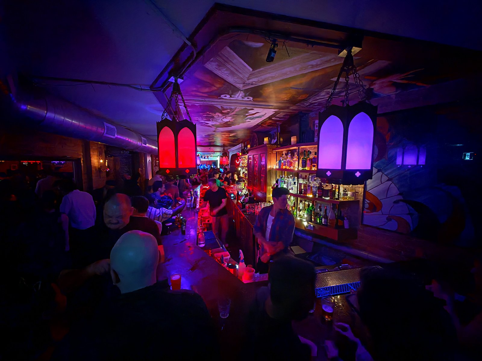 A vibrant, crowded bar at night with colorful lighting, patrons socializing, and a bartender serving drinks under mural-decorated ceilings.
