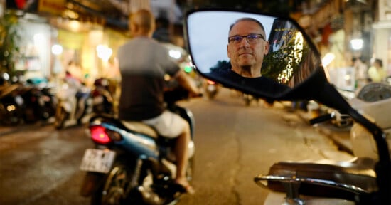 A busy street scene captured at dusk, with a person riding a scooter in the foreground. The reflection of another person wearing glasses is visible in a scooter's rearview mirror, focusing intently. The background is illuminated with various lights and blurred activity.