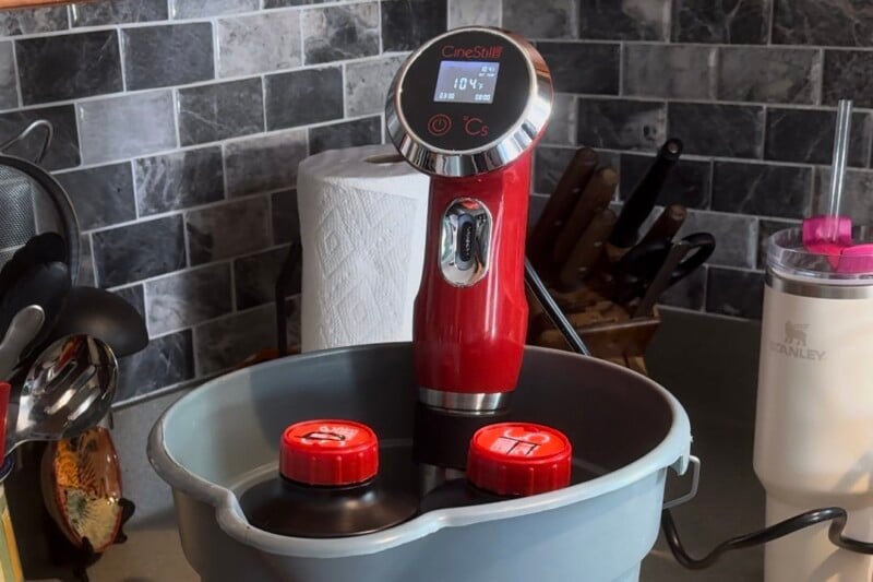 A red immersion circulator with a digital display screen is attached to a bucket filled with two containers. The setup is in a kitchen beside various utensils, a paper towel roll, a knife block, and a cup. The background features a gray tiled backsplash.