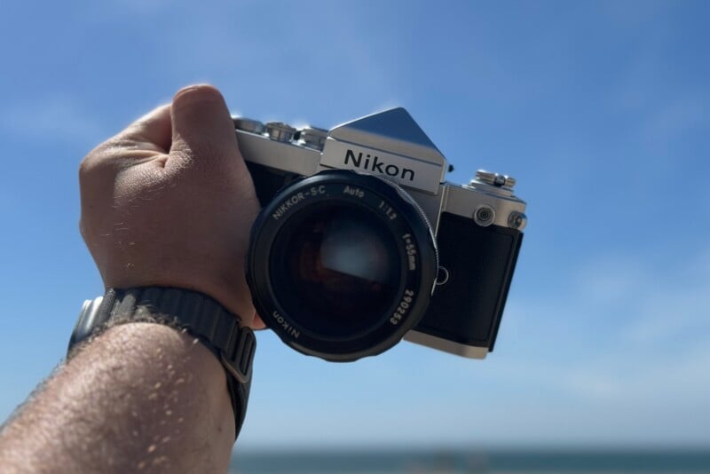 A hand is holding a Nikon film camera under a clear blue sky. The focus is on the camera lens, displaying the Nikkor-S.C Auto 1:1.4 50mm details. A blurred ocean horizon is visible in the background. The person is wearing a watch with a dark strap on their wrist.