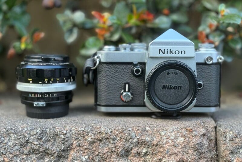 A vintage Nikon film camera with a lens cap sits on a stone ledge next to a detached camera lens. The background features blurred green foliage. The camera has a textured black grip and silver body with "Nikon" branding evident.