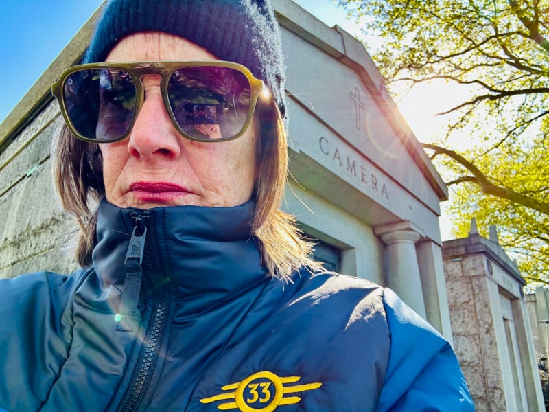 A person wearing a blue jacket with a "33" emblem, dark sunglasses, and a black beanie stands near a stone structure with "CAMERA" inscribed on it. The sun shines through tree branches, creating a bright and contrasting background.