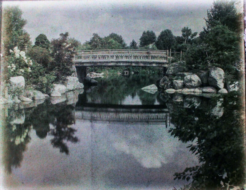 A vintage-style photograph of a serene landscape featuring a stone bridge arching over a calm river, flanked by lush trees and large rocks, with reflections visible in the water.