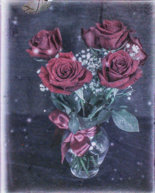 A vintage-style image of a bouquet of deep red roses in a glass vase, with small white flowers and green leaves, set against a textured dark blue backdrop with light speckles.