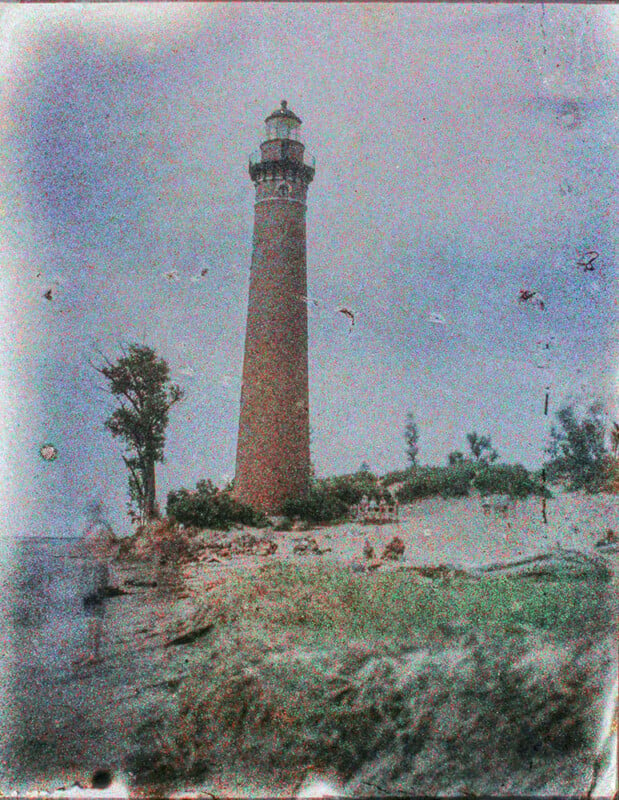 An aged, grainy photo of a tall, reddish-brown lighthouse standing amidst lush greenery, with a cloudy sky overhead and blurred spots on the image, possibly from water drops or photo aging.