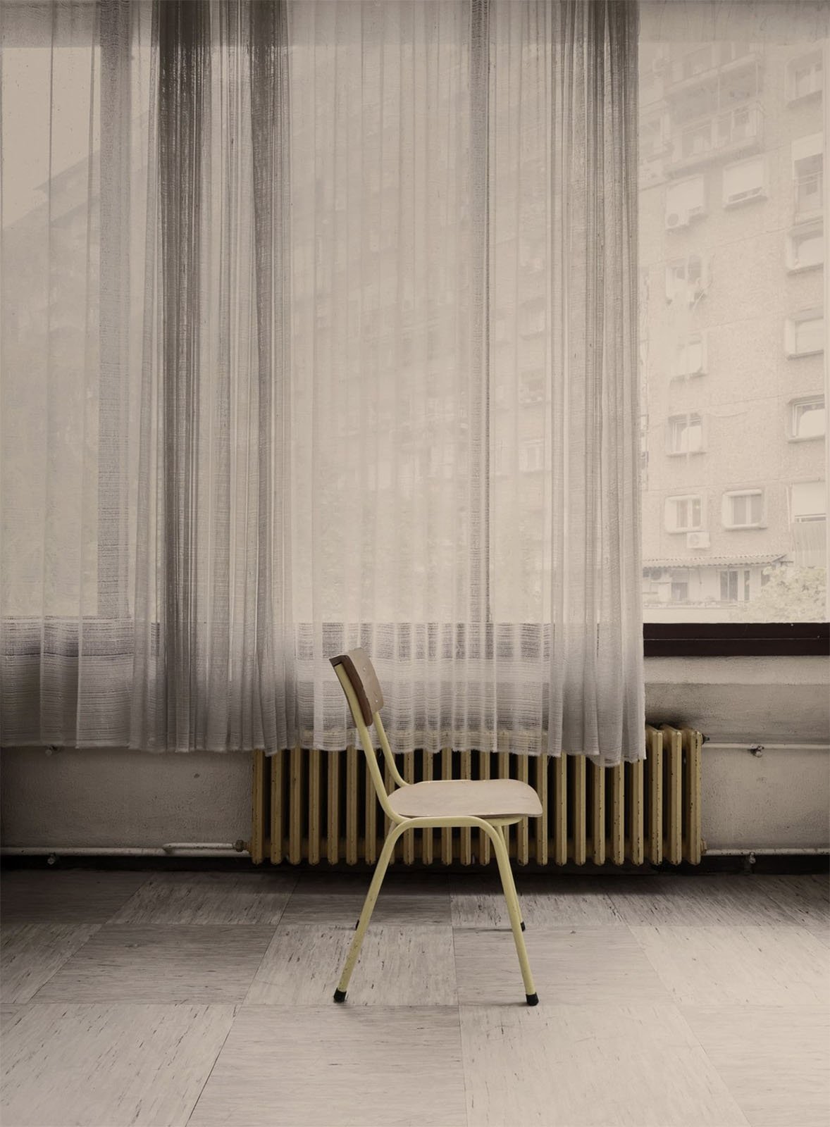 A single vintage chair stands in front of a window draped with sheer curtains, overlooking a cityscape, emphasizing a quiet, melancholic atmosphere in a room with a wooden floor.