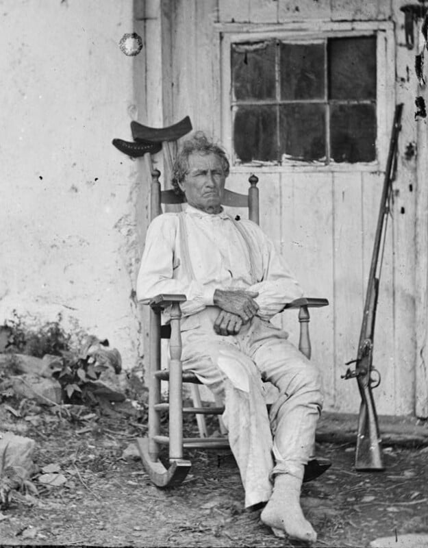 An older man with disheveled hair is sitting in a wooden rocking chair in front of a weathered door. He is wearing a light-colored shirt and pants, and is barefoot. A rifle is propped up against the wall beside him. The setting appears to be rustic and outdoors.