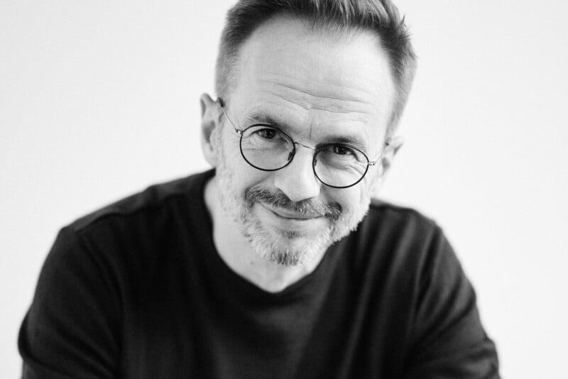 Black and white portrait of a smiling middle-aged man with glasses, sporting a beard and wearing a black t-shirt, against a plain background.