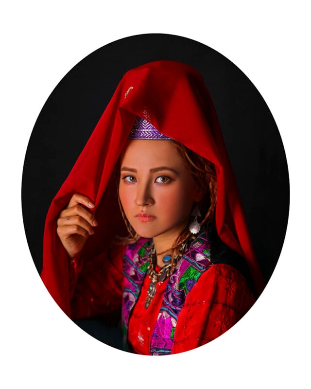 A young person wearing traditional colorful clothing and jewelry, with a red headscarf partially covering their head. The individual is set against a dark background and gazes directly at the camera, holding the edge of the headscarf with one hand.