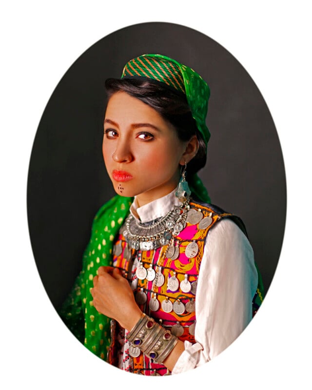 A woman stands against a dark background, dressed in traditional, colorful attire. She wears a vibrant, multicolored vest adorned with coins, a green headscarf with gold patterns, large silver earrings, and intricate silver jewelry on her chest and wrist.