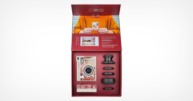 A red box is open displaying a camera kit. The kit includes a vintage-style camera, several detachable lenses, and other accessories. The inside cover of the box features a person in an orange sweater holding a developed photo with additional text and images.