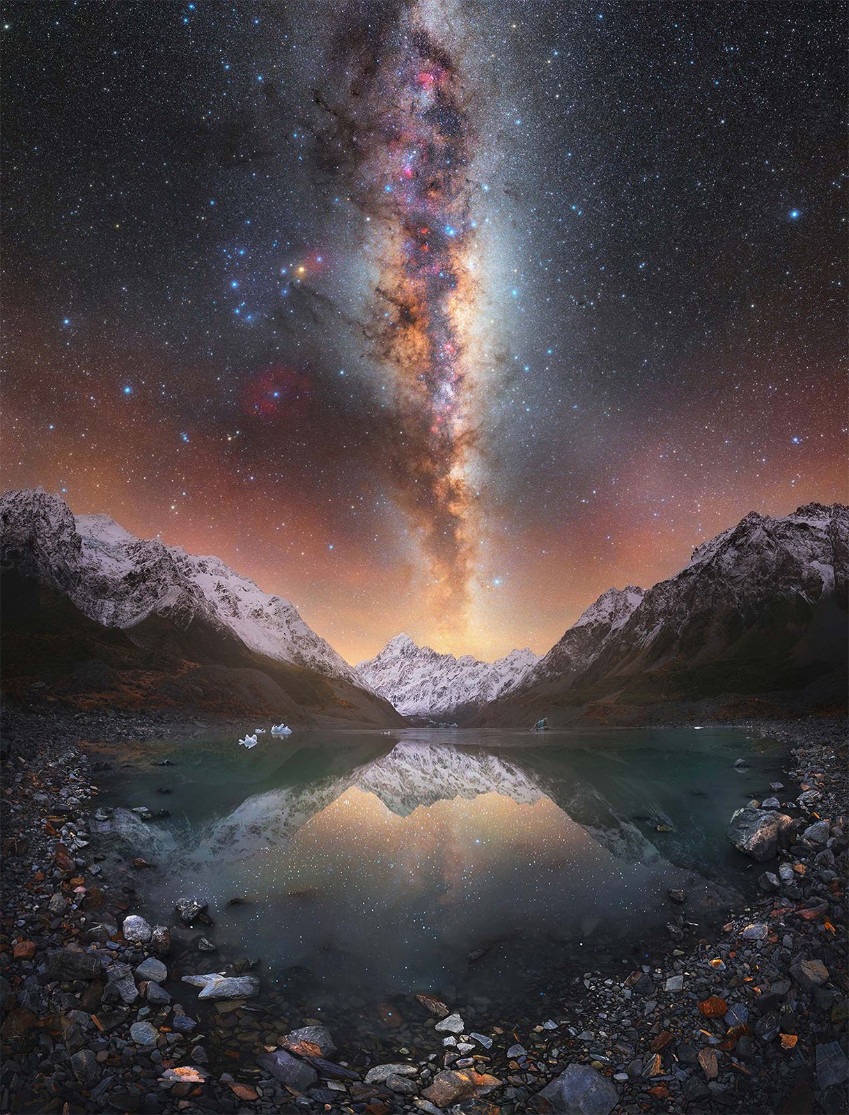 A stunning night sky filled with stars and the Milky Way stretches vertically over snow-capped mountains that are reflected in a clear, calm lake below. The scene is serene, with rocky terrain in the foreground leading to the tranquil waters and majestic peaks.
