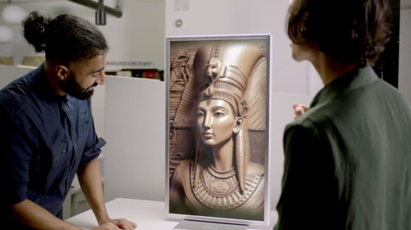 Two people, one male and one female, examine a digital artwork of an ancient Egyptian queen displayed on a monitor in a modern studio setting.