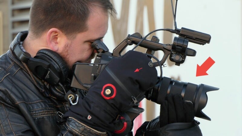 A person is operating a professional video camera while wearing a leather jacket and black gloves. The individual is also wearing large over-ear headphones. A red arrow is pointing to a component on the camera, highlighting a detail.