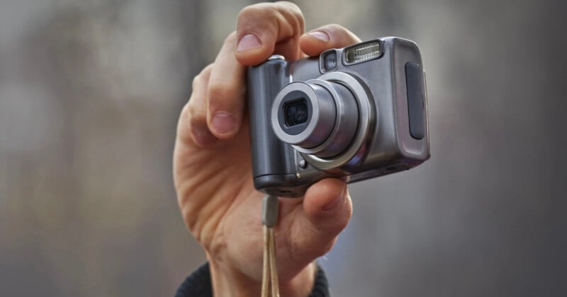 A close-up of a hand holding a silver compact digital camera with the lens extended, ready for taking a photo. The background is blurred, making the camera and hand the focal points of the image.