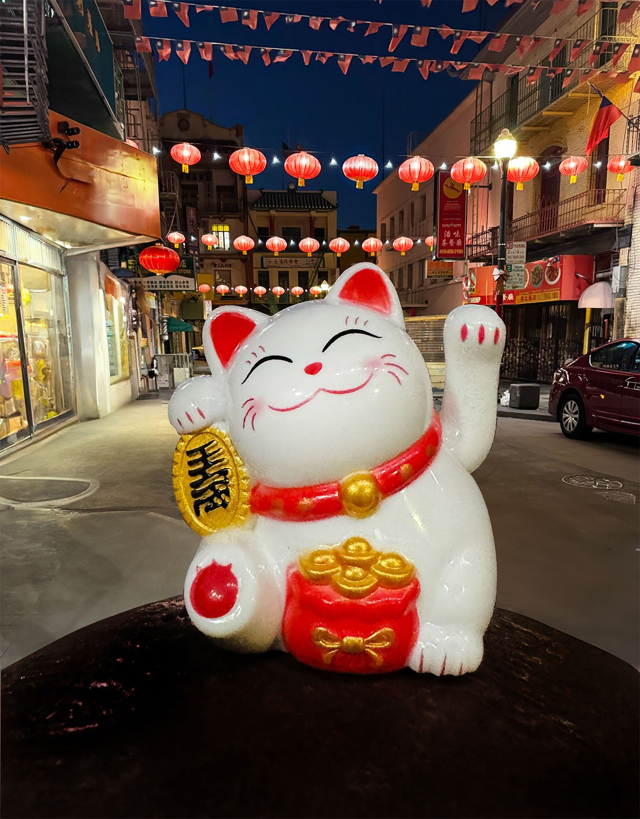 A white and red Maneki-neko (beckoning cat) figurine with a raised paw and golden accents is prominently displayed in the foreground. In the background, there is a street adorned with red lanterns and warmly lit shops at dusk.