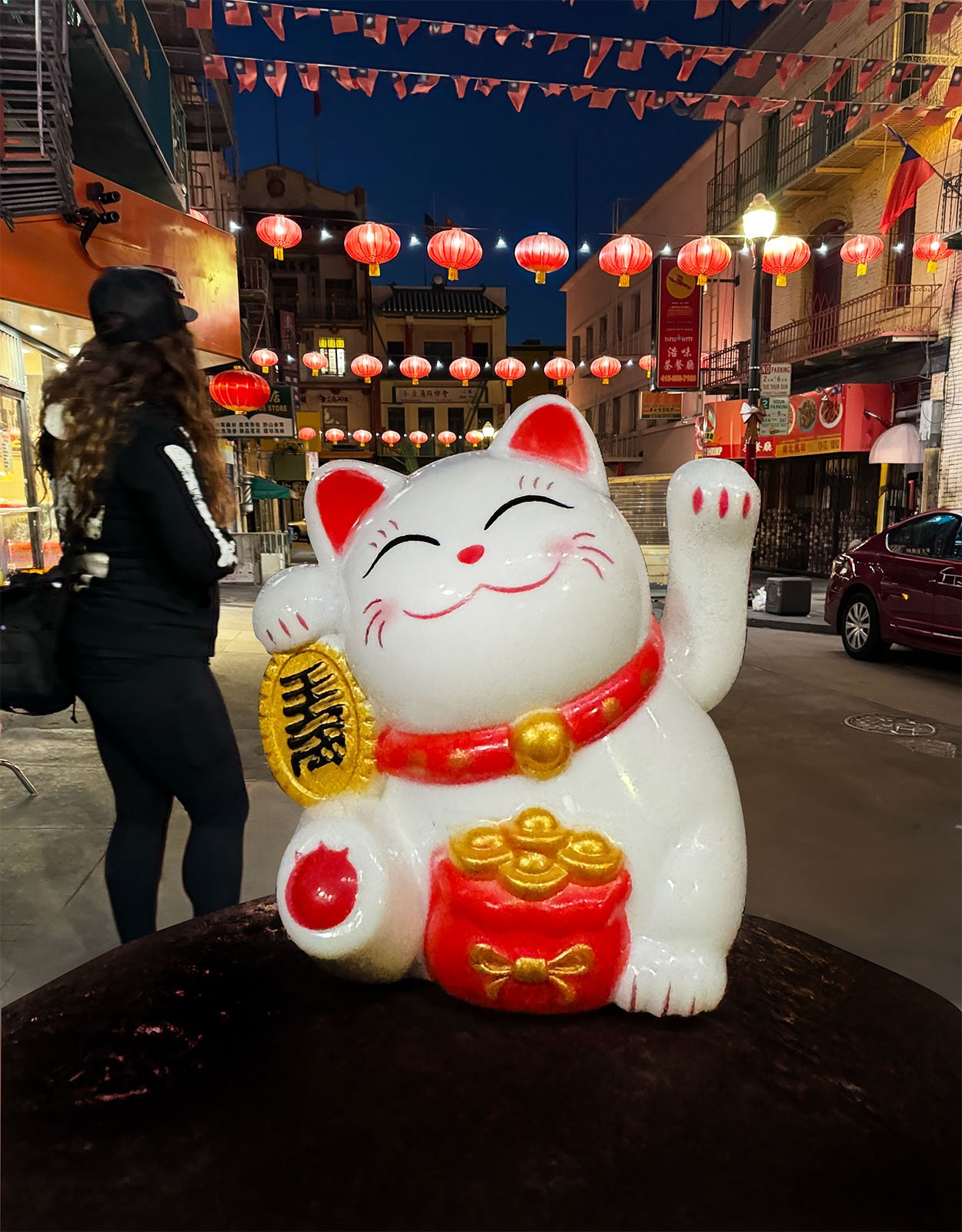 A large Maneki-neko (beckoning cat) statue with a raised paw and a red collar stands prominently in a lantern-lit street. In the background, a person wearing a black outfit and cap walks away, and red lanterns hang overhead, illuminating the evening scene.