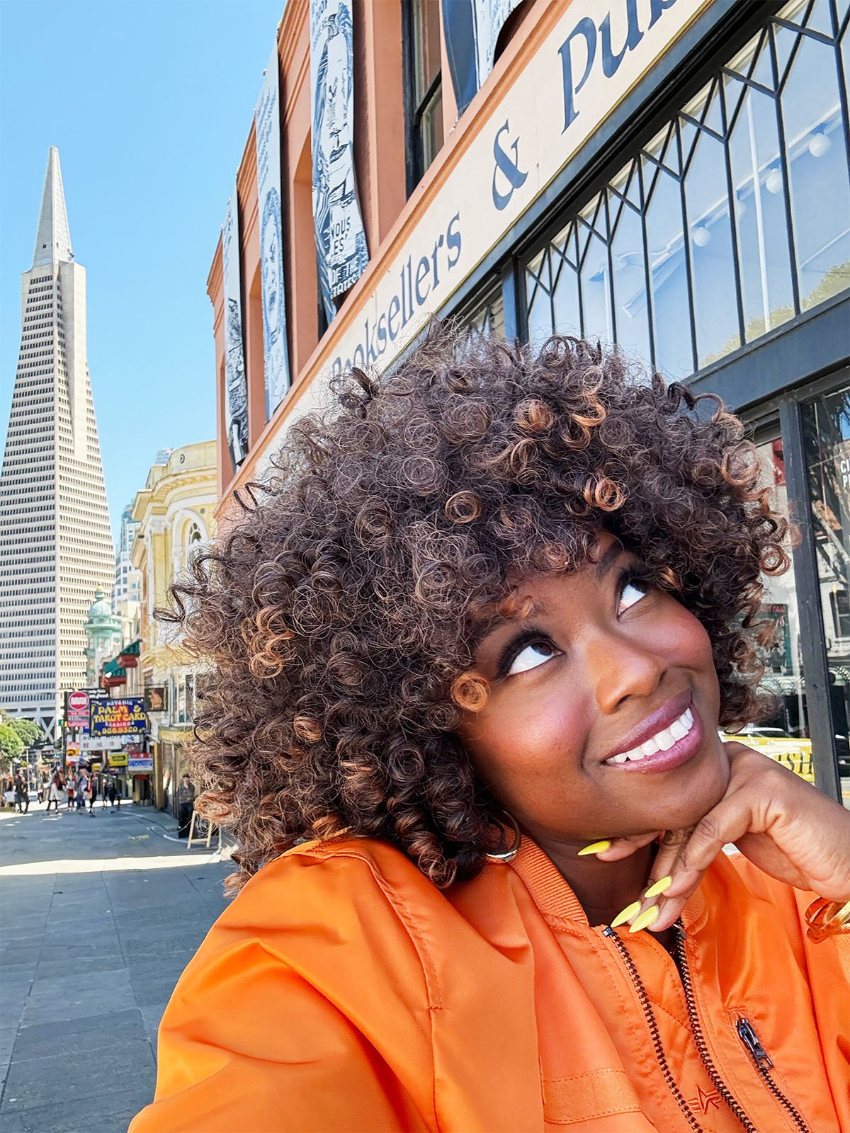 A woman with curly hair and an orange jacket smiles while looking upwards. She is standing on a city street with a tall, pointed skyscraper in the background. The façade of a bookstore is also visible.