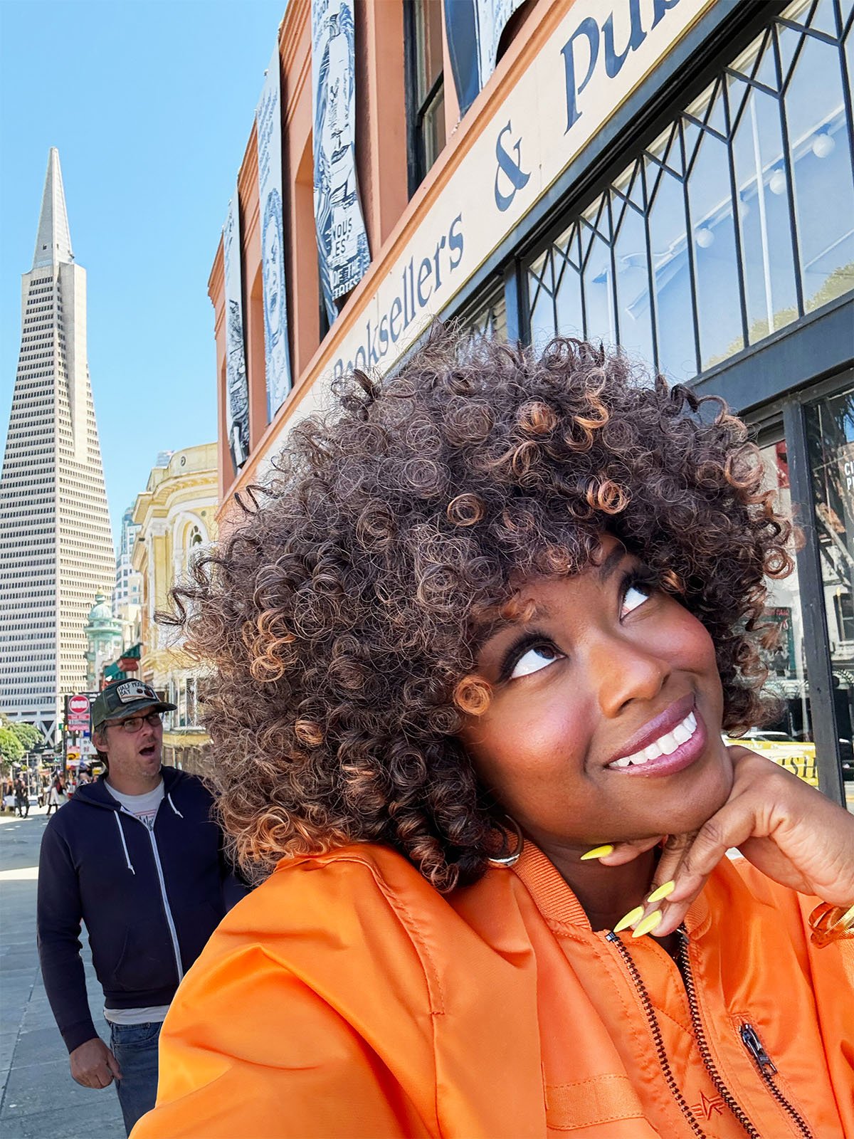 A woman with curly hair and an orange jacket smiles while looking up, standing in front of a building with a sign that reads "Booksellers & Publishers." A man in a hoodie and cap appears surprised in the background. The Transamerica Pyramid is visible behind them.