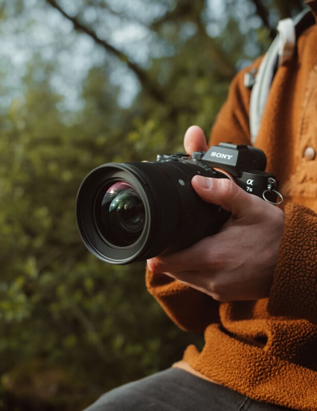 Person wearing a rust-colored jacket holds a Sony Alpha 7R camera outdoors with a blurred natural background. The person's face is not visible, and the focus is on the camera.