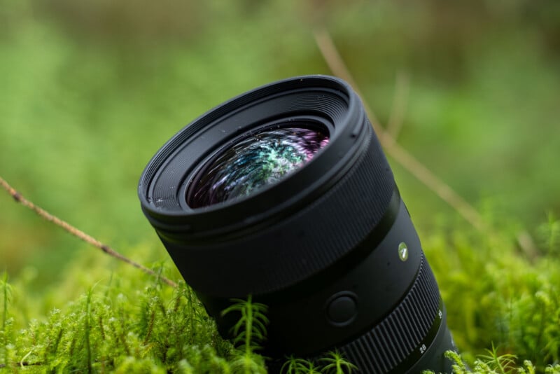 A black camera lens lying on a bed of moss in a natural outdoor setting. The lens is angled slightly, showing its reflective glass elements with green and purple hues. The background is blurred, highlighting the lens as the focal point of the image.