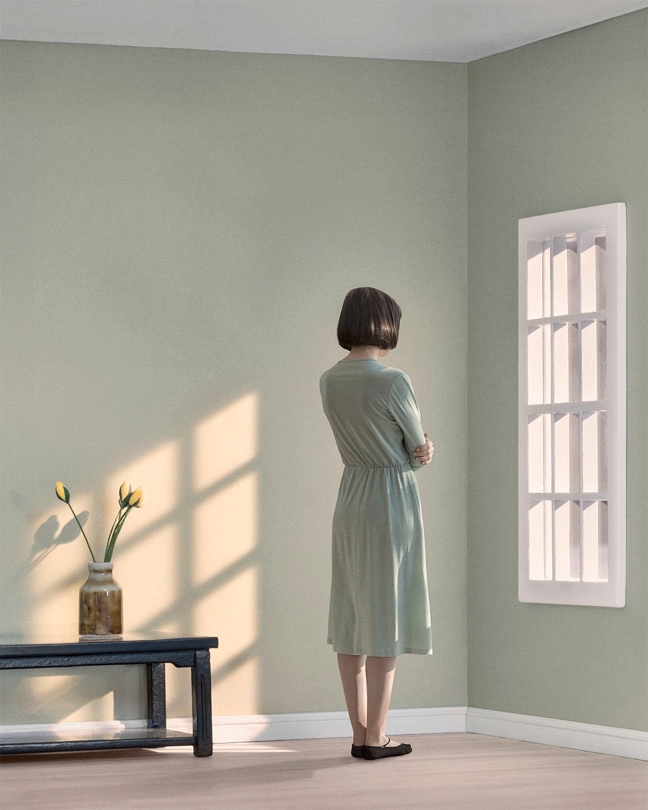 A woman in a teal dress stands facing a wall in a minimalistic room, gazing at a window with light casting shadows on the wall. a small table with a vase and flowers is nearby.
