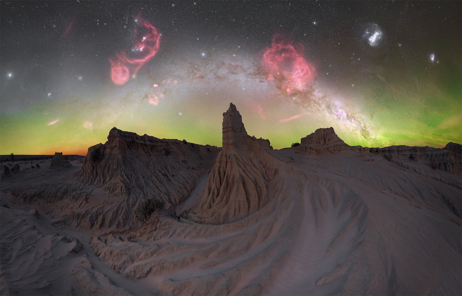 A breathtaking night-time landscape of a desert with prominent rock formations in the foreground. The sky features a stunning display of stars, vibrant pink and red nebulae, and faint green auroras illuminating the horizon, creating a surreal celestial scene.