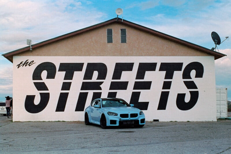 A light blue sports car is parked in front of a beige building with large, bold black letters reading "THE STREETS" painted on the side. The background features a partly cloudy sky, and the scene appears to be in an open area or parking lot.