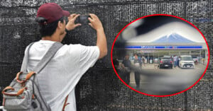 A person in a red cap and white shirt is taking a photo with a smartphone. They are standing in front of a black mesh wall. A circular inset shows a Lawson convenience store with Mount Fuji visible in the background.