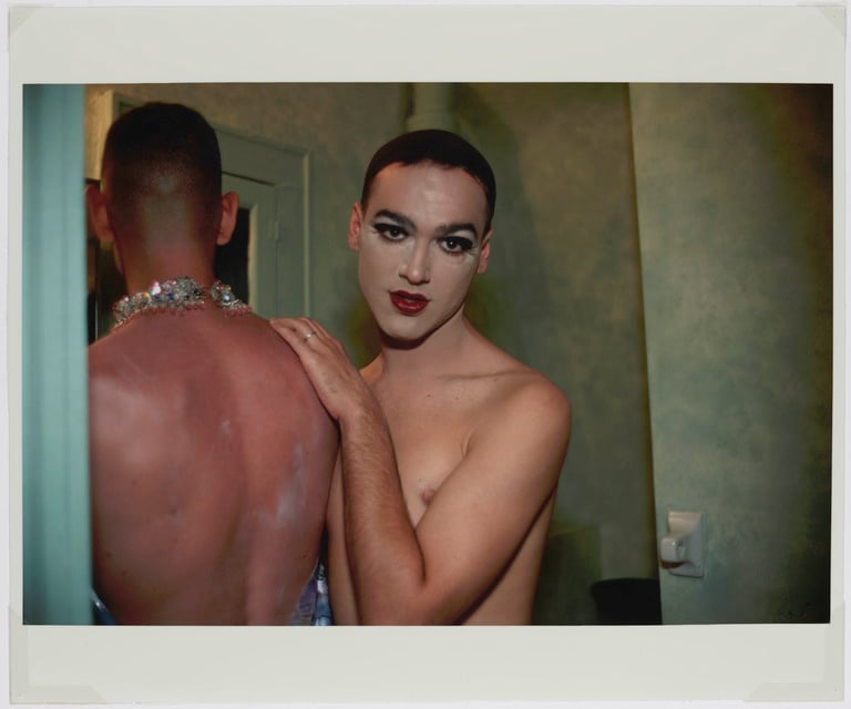 Two shirtless individuals are standing close together in a dimly lit room. One faces away, showing a red, sunburnt back and sparkly jewelry. The other faces the camera, wearing makeup with bold eyeshadow and red lipstick, and has a hand resting on the other's shoulder.