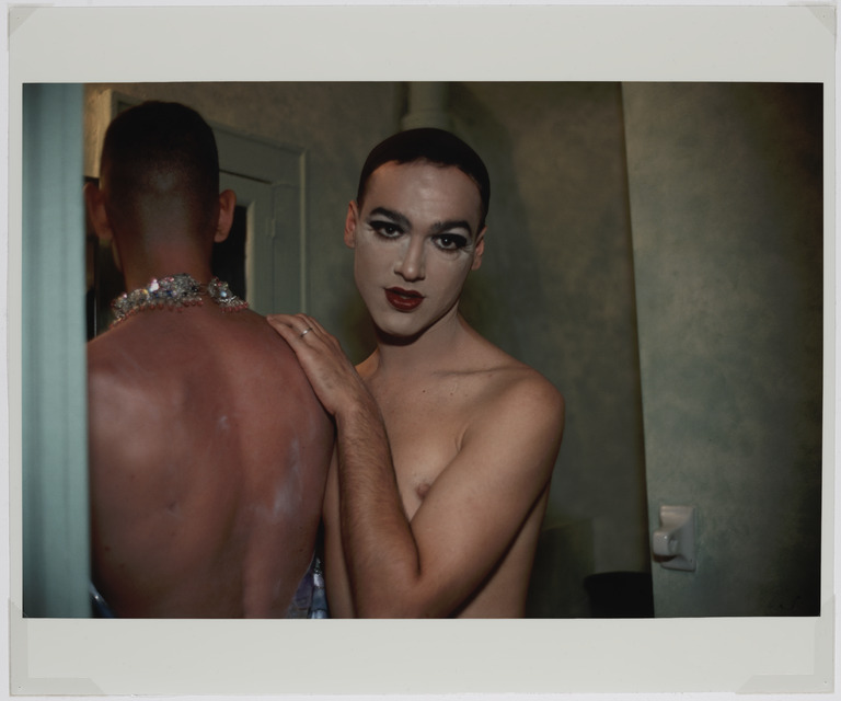 Two shirtless individuals are standing close together in a dimly lit room. One faces away, showing a red, sunburnt back and sparkly jewelry. The other faces the camera, wearing makeup with bold eyeshadow and red lipstick, and has a hand resting on the other's shoulder.