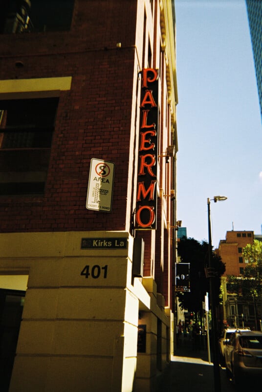 A red-brick building with a neon sign that reads "PALERMO" vertically hangs from the side. The building is located at the corner of Kirks Lane and another street, as indicated by the street sign. Cars are parked along the street in a sunny, urban setting.