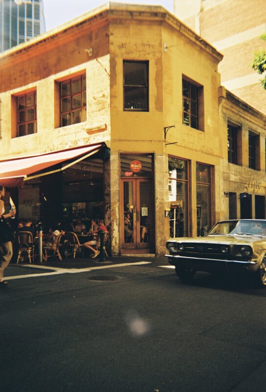 A two-story building on a sunny street corner houses a café with some outdoor seating. A classic car drives by in the foreground. The building has a weathered, yellow facade with red-framed windows, and a sign above the entrance reads "Maisy's Café.