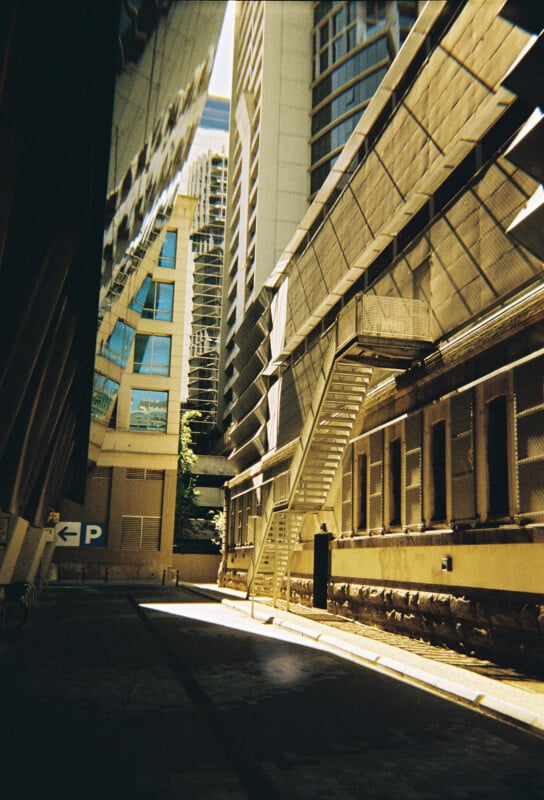 An urban alleyway framed by modern high-rise buildings. The scene features a mix of glass and concrete architecture with sunlight streaming through, casting shadows. A metal staircase is attached to one building, and a "P" parking sign is visible on the left.