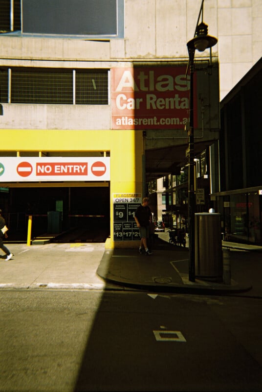 A parking structure entrance with a yellow and red "No Entry" sign. A billboard above advertises "Atlas Car Rental." A person stands near the entrance. A sign nearby shows parking rates and mentions "Open 24/7." The structure is in an urban setting.