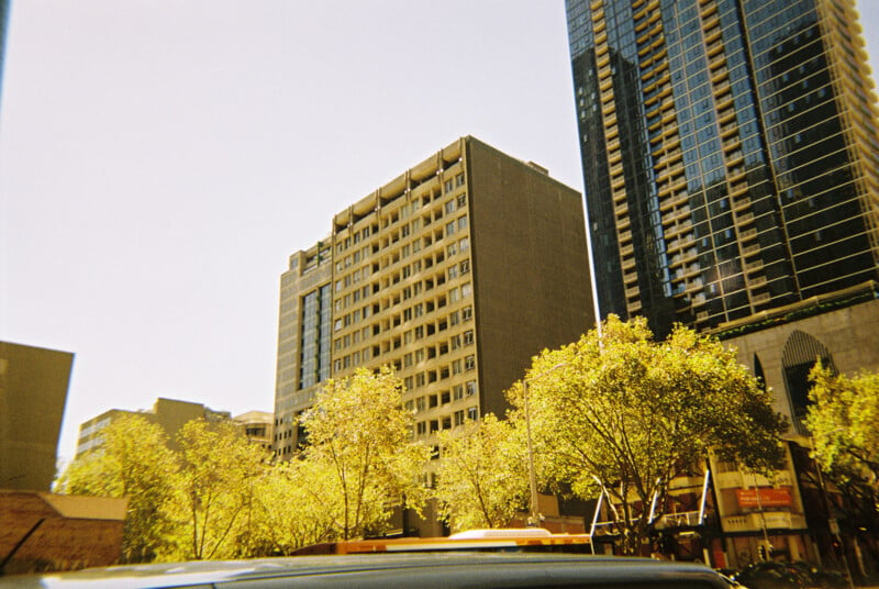 A sunny cityscape featuring several high-rise buildings. The image shows a mix of older and newer architecture, with trees in the foreground adding greenery. The sky is clear and blue, casting a warm light on the scene.