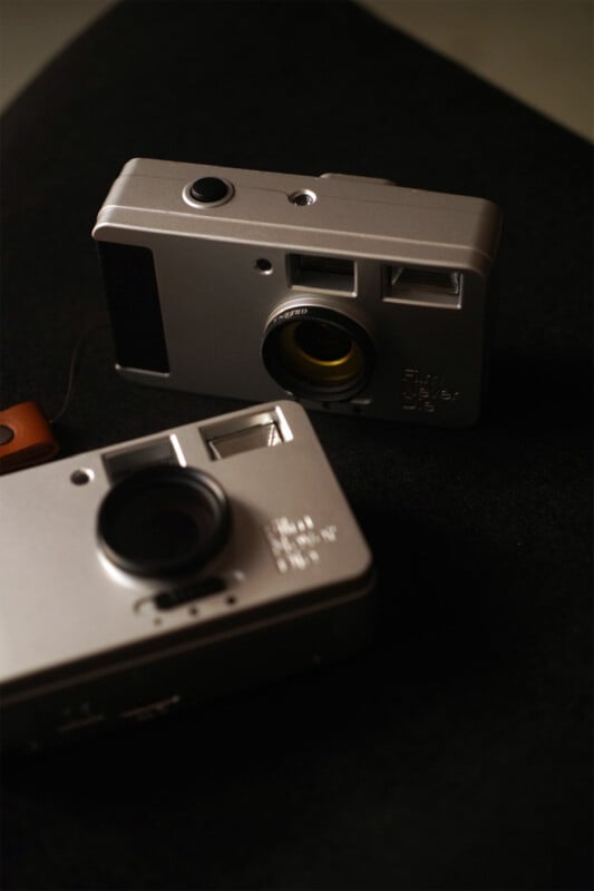 Two vintage-style silver cameras are placed on a dark surface. The camera in the foreground has a lens pointing upwards, while the camera in the background has its lens facing forward, emphasizing its yellow inner ring.