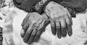 Close-up black-and-white photo of elderly hands resting on a white cloth. The hands show prominent veins and wrinkles, with fingernails trimmed short. The person appears to be wearing a garment with intricate embroidered patterns on the sleeves.